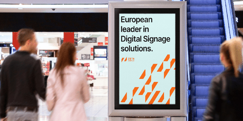 ZetaDisplay digital signage screen in a shopping mall reading 'European leader in Digital Signage solutions,' with shoppers passing by near an escalator.